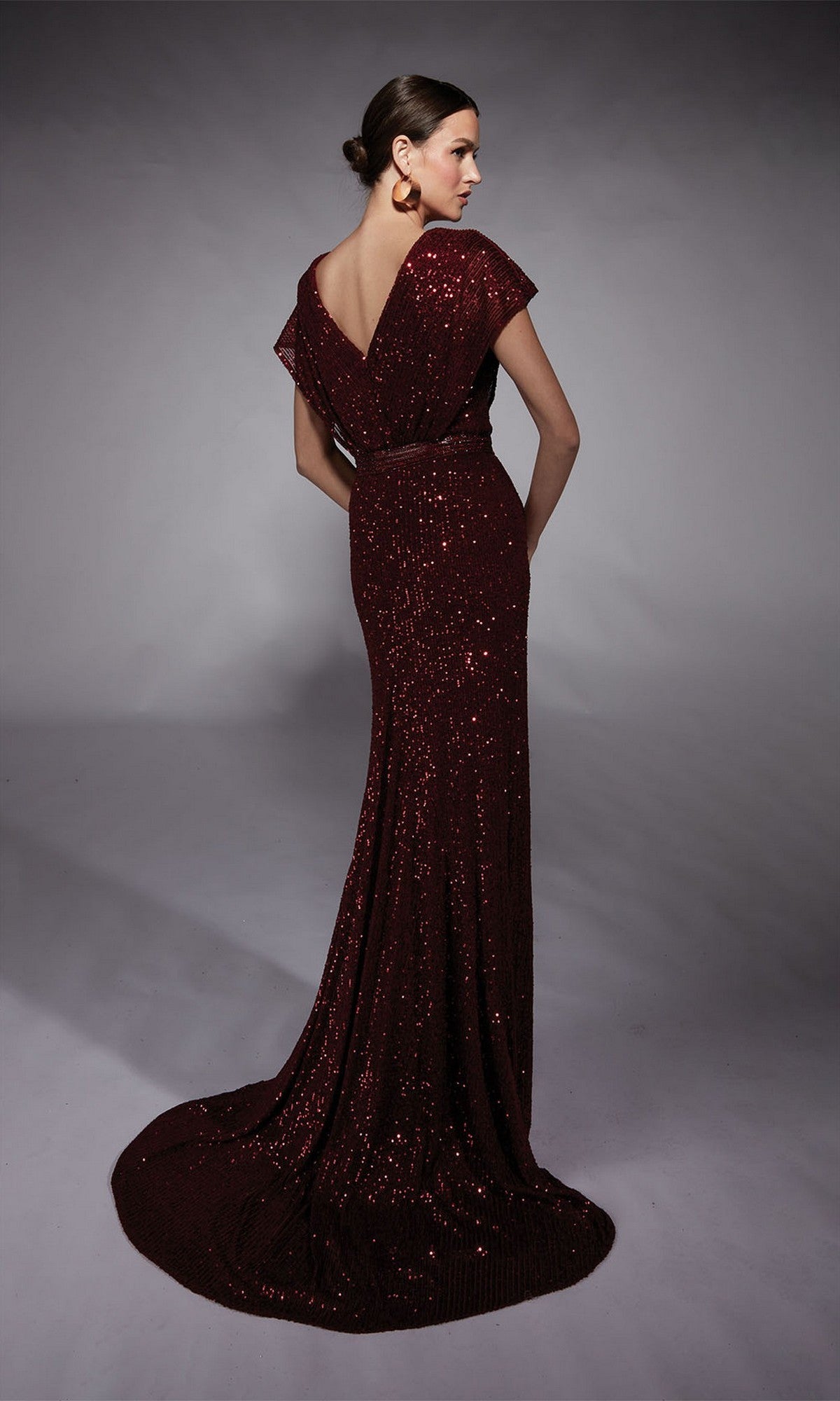 A woman stands on a grey backdrop wearing the Alyce 27708 long formal dress, a dark red sequin evening gown featuring a deep V-back, cap sleeves, and a beaded waistband. Her hair is styled in a low bun.